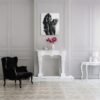 Black-and-white figurative painting of an embrace displayed above a white fireplace in a classic, elegant interior with a black armchair and floral decor.