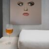 Modern face art painting of a surreal woman’s portrait displayed against a textured wall with a glass of wine and a white cushion.