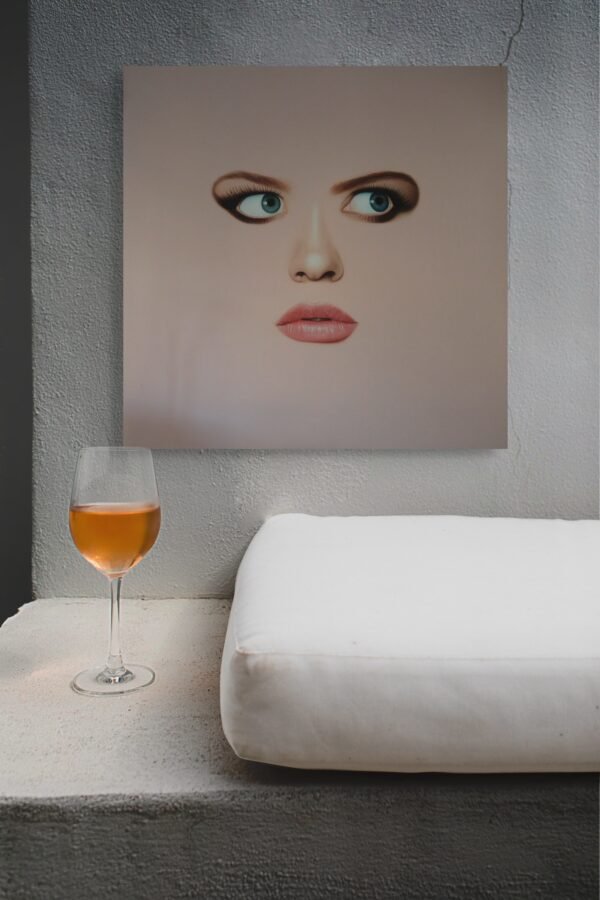 Modern face art painting of a surreal woman’s portrait displayed against a textured wall with a glass of wine and a white cushion.